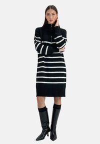 Gestreifter schwarz-weißer Pulloverkleid mit einem Reißverschluss-Hochkragen. Gerippte Bündchen und Saum, präsentiert mit knielangen schwarzen Stiefeln mit Absatz.