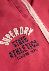 Rød sweatshirt med "SUPERDRY" og "STATE ATHLETICS" broderi i hvid og navy. Teksturen er blød med designdetalje af halv lynlås.