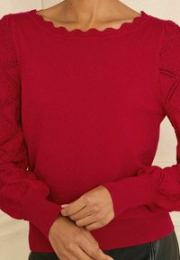 Punainen neulepusero, jossa on aaltoileva kaulus ja teksturoitu pitsikuvioitu hihansuu. Tiukat hihansuut ja helma, pehmeä materiaali, jossa on ribbireunukset.