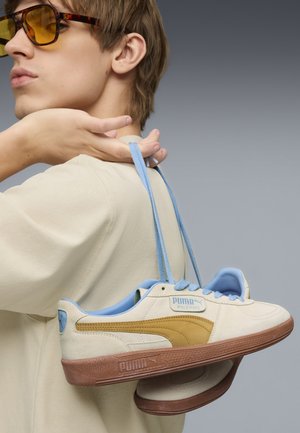 Puma-Sneaker mit einer cremefarbenen Oberseite, blauen Akzenten, brauner Sohle und strukturiertem Material, gehalten von einer Person mit hellblauen Schnürsenkeln, die herunterhängen.