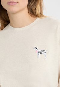 Jasnobeżowa bluza z małym, haftowanym dalmatyńczykiem z czarnymi plamami i różowym szalikiem na szyi, umieszczonym na lewej stronie klatki piersiowej.