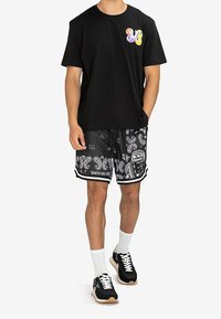 Zwart katoenen T-shirt met een kleurrijke grafische print op de borst, gecombineerd met zwarte shorts met paisley-patroon en zwarte sneakers met witte accenten.