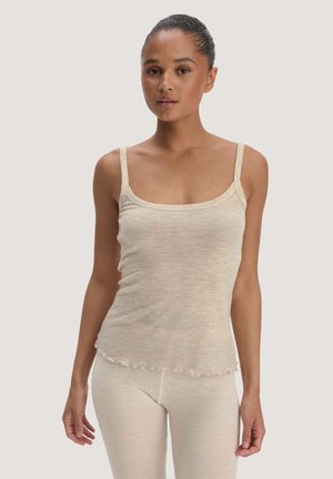 Frau mit Haaren zu einem Dutt zusammengebunden, trägt ein beiges ärmelloses Tanktop und passende Hose und steht vor einem schlichten hellen Hintergrund.
