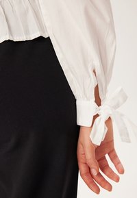 Weiße Bluse mit Puffärmeln, ausgestattet mit elastischen Bündchen, die mit einem weißen Band gebunden sind. Sichtbare glatte Textur und leichter Glanz vor schwarzem Hintergrund.