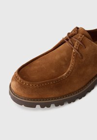 Scarpa in suede marrone con una texture liscia, punta rotonda e lacci marroni. Presenta una suola in gomma spessa e robusta con un battistrada rigato per una buona aderenza.