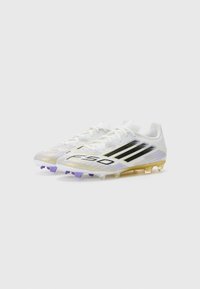 Des chaussures de football blanches avec des rayures noires, présentant un design en dégradé texturé, des accents violet et or, et une silhouette basse.