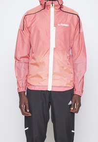 Leichter Windbreaker in rot-weißen Streifen mit Kapuze, Frontreißverschluss und Brusttasche, kombiniert mit schwarzen Hosen mit weißen Akzenten.