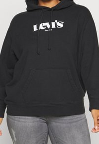 Persona che indossa un hoodie nero di Levi's con testo del logo bianco e jeans grigi, in piedi davanti a uno sfondo neutro.