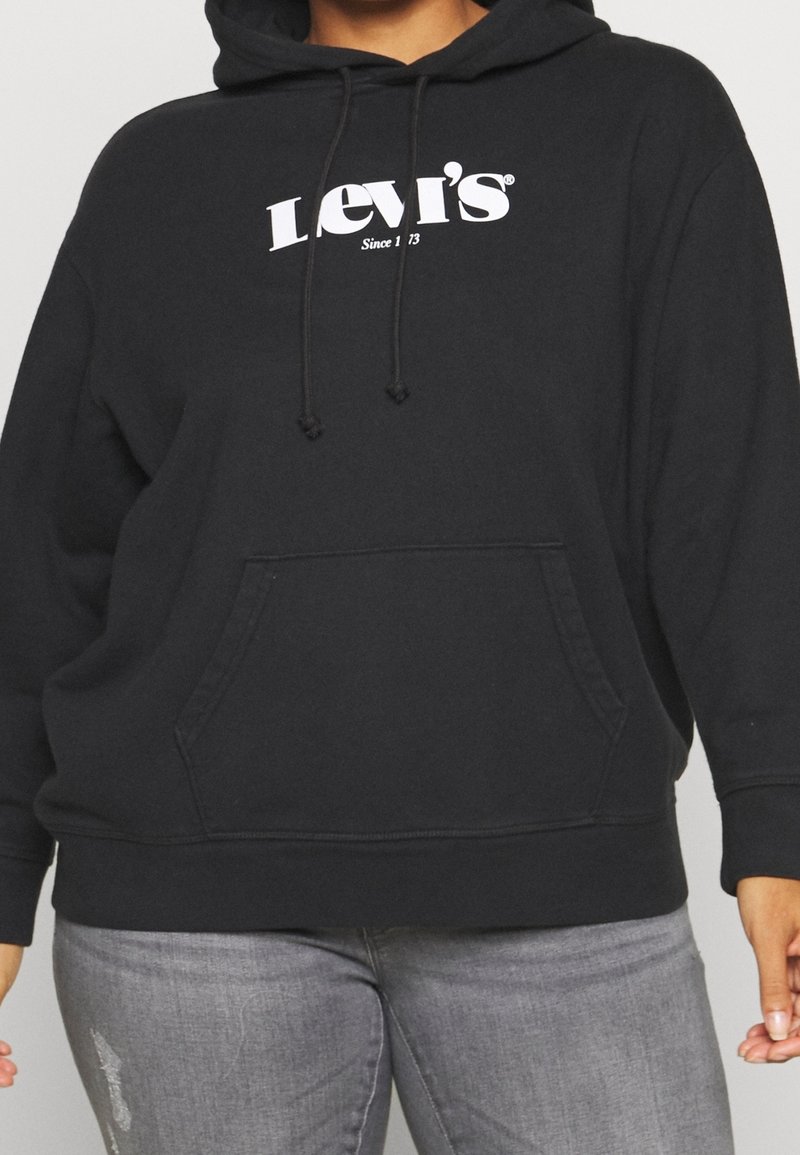 Persona che indossa un hoodie nero di Levi's con testo del logo bianco e jeans grigi, in piedi davanti a uno sfondo neutro.
