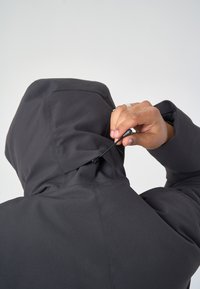 Schwarz gewandete Kapuzenjacke mit glatter Textur; verstellbarer Kordelzug, der am Rücken der Kapuze festgezogen wird; minimalistisches Design und funktionale Details.