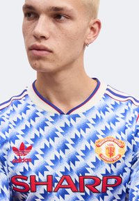 Jeune homme aux cheveux blonds courts et portant une boucle d’oreille en anneau, vêtu d’un maillot de football de Manchester United à motif zigzag bleu et blanc.