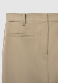 Gros plan sur un pantalon beige montrant la taille avec des passants de ceinture et une poche fendue sur le côté droit.