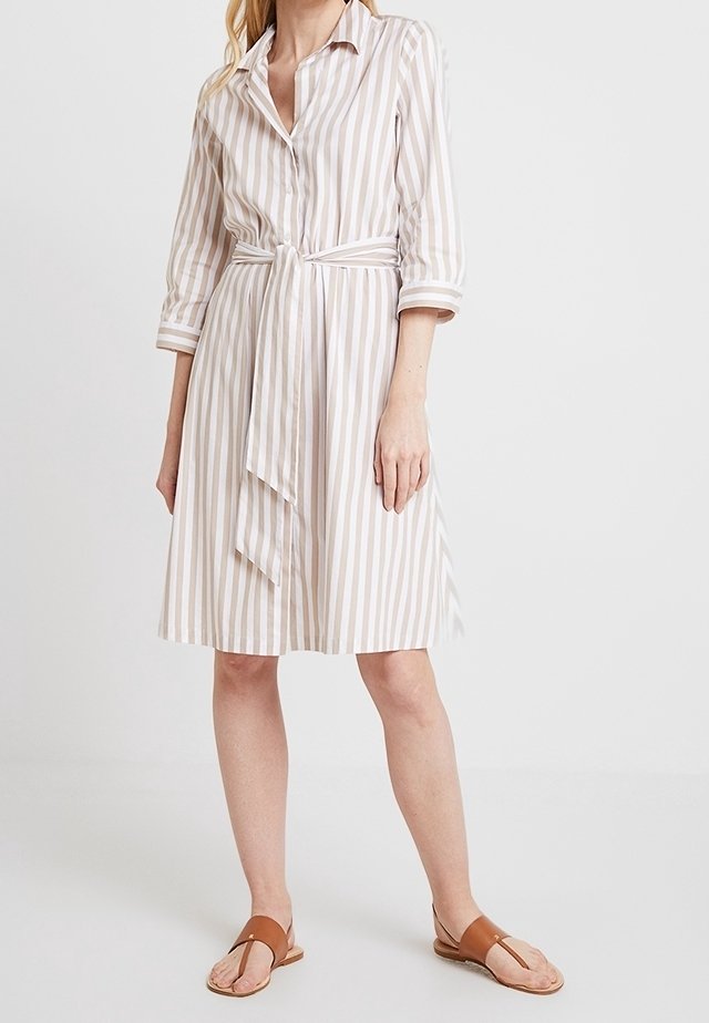 Femme portant une robe-chemise à rayures verticales beige et blanc avec des manches trois-quarts et une ceinture à la taille, associée à des sandales plates marron.