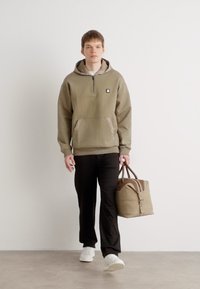 Sudadera con cierre en color beige, con bolsillo frontal y logo, combinada con pantalones negros y zapatillas blancas. Bolsa marrón con asas de dos tonos.