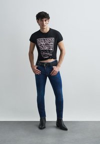 Camiseta recortada negra con estampado gráfico, jeans ajustados azules, cinturón negro con detalles circulares y botines negros sobre fondo gris.