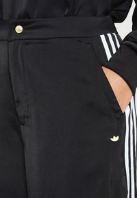 Czarne spodnie sportowe z białymi paskami po bokach, małym złotym logo obok kieszeni oraz złotym guzikiem na pasku, ręka w kieszeni.