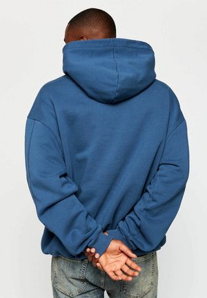 Hombre con sudadera azul con capucha y vaqueros de pie de espaldas a la cámara, con las manos entrelazadas detrás de la espalda contra un fondo blanco liso.