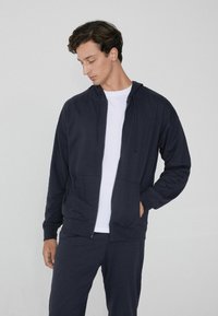 Sudadera con cremallera en color azul marino de tejido suave, con un bolsillo delantero, capucha con cordón y un corte relajado, combinada con una camiseta blanca debajo.