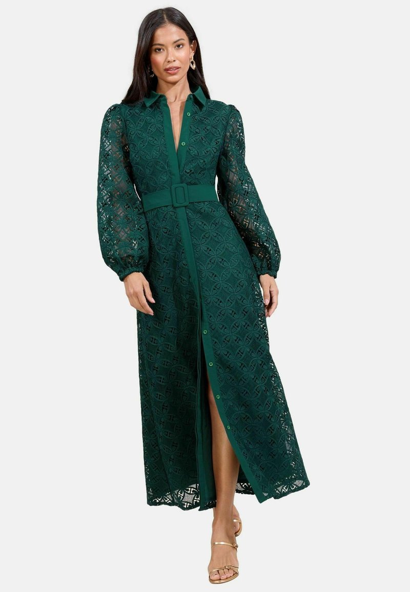 Maxi abito verde in pizzo con chiusura a bottoni sul davanti, vita cintrata con cintura e maniche a sbuffo, caratterizzato da un motivo geometrico e uno spacco su un lato.