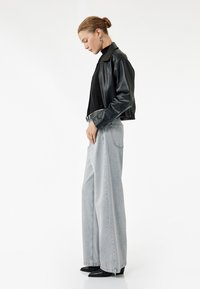 Chaqueta corta de cuero sintético negra sobre un cuello de tortuga negro, combinada con jeans de pierna ancha de talle alto en gris claro y botas de tacón puntiagudas negras.