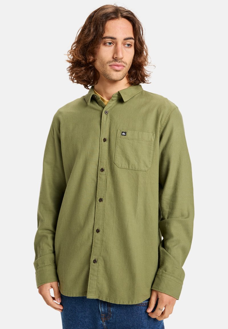 Camisa de franela verde con cuello abotonado, bolsillo frontal y botones oscuros. Textura suave con un corte relajado y mangas largas.