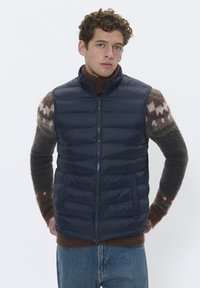 Marineblauer Steppweste mit einem Reißverschluss, zwei Seitentaschen, gestepptem Design, getragen über einem gemusterten, gestrickten Pullover, kombiniert mit blauen Jeans.