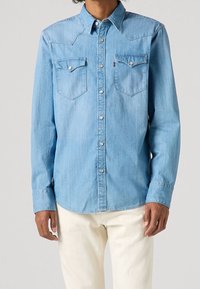 Vaaleansininen farkkupaita, jossa on kaulus, paineknopit ja kaksi rintataskua. Varustettu länkkärityylisellä selkäkopilla ja pitkillä hihoilla.