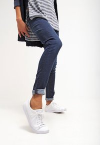 Vita canvas sneakers med runda tår och platta sulor, som bärs med mörkblå uppvikta jeans och en randig grå topp, framför en vit bakgrund.