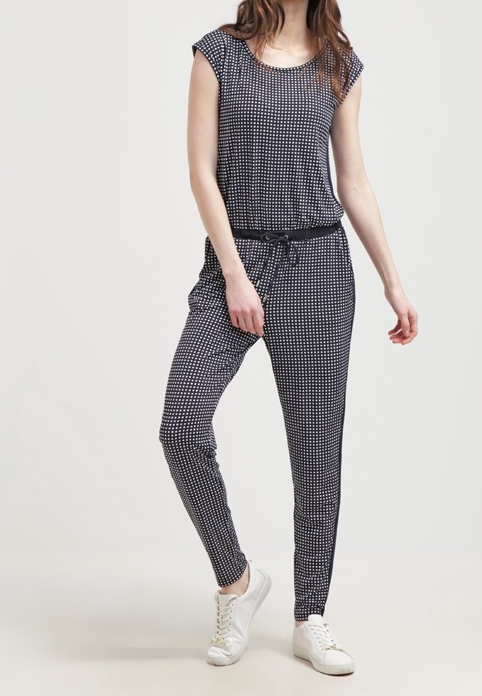 Femme portant une combinaison noire et blanche à petits carreaux avec une ceinture noire à cordon et des baskets blanches, debout devant un fond uni.