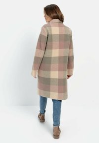 Lang geruite jas van zacht materiaal in tinten roze, beige en groen. Het heeft een kraag en is gecombineerd met lichtblauwe jeans en bruine laarzen.