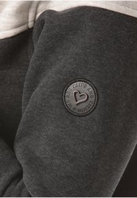 Sudadera gris oscuro con capucha gris claro, tela texturizada y una etiqueta circular negra con un diseño de corazón en relieve en la manga.