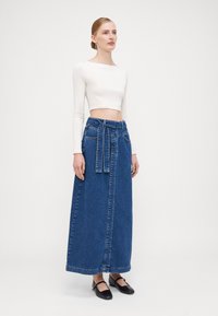 Lang blå denimskjørt med knyttet midje, har fem lommer og rett kutt. Kombinert med en tettsittende hvit langermet topp og svarte sko.