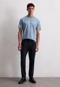 Jonge man die staat, gekleed in een lichtblauw Marc O'Polo t-shirt, zwarte broek en zwarte leren schoenen tegen een effen witte muur.