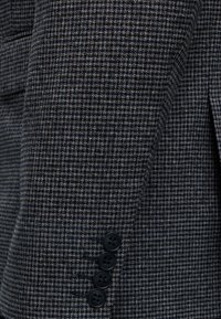 Manche de blazer à motif pied-de-poule gris foncé et noir avec quatre boutons noirs en rangée verticale près du poignet.