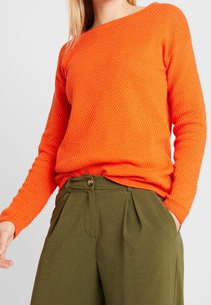 Femme portant un pull à manches longues texturé orange vif, rentré dans un pantalon large taille haute vert olive avec bouton et plis.