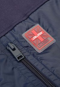 Marine nylon jas met een zwarte rits en een rood en grijs rubberen logo patch met een Union Jack ontwerp. Getextureerde, gevoerde stof.