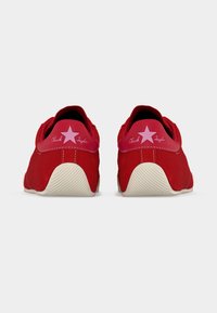 Ζευγάρι κόκκινων χαμηλών αθλητικών παπουτσιών Chuck Taylor, εμφανίζεται από πίσω με ροζ λογότυπα αστεριών και λευκές λαστιχένιες σόλες.
