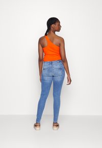 Top de alça única laranja combinado com calças skinny azuis claras e sandálias bege, exibindo um design ajustado e uma textura de tecido suave.