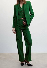 Femme debout portant un tailleur trois pièces vert avec blazer, gilet boutonné, pantalon assorti et escarpins noirs à bout pointu sur un fond neutre.