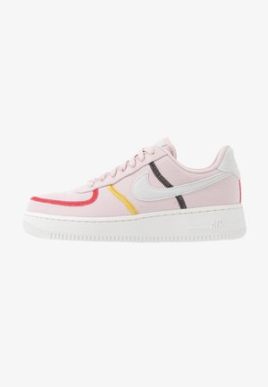 Baskets basses roses avec semelle blanche, swoosh Nike blanc, et détails rouges, jaunes et noirs cousus sur le côté.