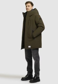 Green padded coat with a high collar, side pockets, and black accents. Paired with dark gray jeans and black lace-up boots.