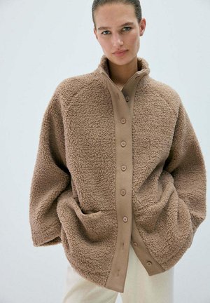 Mujer con chaqueta de polar marrón de gran tamaño con botones y grandes bolsillos, de pie frente a un fondo claro y liso.