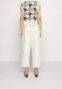 Jasnoszary top bez rękawów z geometrycznym brązowym wzorem, zestawiony z luźnymi, kremowymi spodniami o szerokich nogawkach oraz czarnymi sandałami na obcasie.