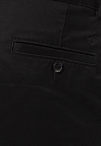 Pantaloni neri in cotone con una tasca frontale singola e chiusura a bottone, caratterizzati da un delicato dettaglio di cuciture che conferisce una texture liscia.