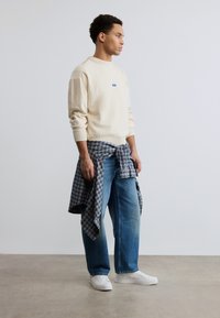 Homme portant un pull crème, un jean bleu, des baskets blanches et une chemise grise à carreaux nouée autour de la taille, debout devant un mur blanc uni.