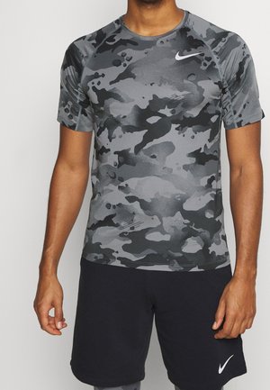 Mężczyzna w szarej i czarnej koszulce sportowej z krótkim rękawem w kamuflaż oraz czarnych szortach z białymi logo Nike na piersi i nodze.