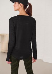 Schwarzes Langarmshirt aus leichtem, strukturiertem Stoff. Kombiniert mit dunkelgrünen Camouflage-Leggings im entspannten Schnitt und mit Raglanärmeln.