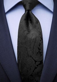 Svart slips med paisleymönster i slätt tyg, bärs över en ljusblå skjorta och mörk kostymjacka. Mönstret är subtilt och strukturerat.
