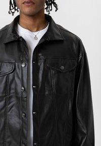 Zwarte leren jas met knoopsluiting, grote kraag, twee borstzakken en een gladde textuur. Draagt over een wit T-shirt.