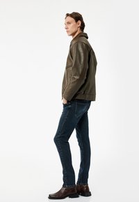 Olivgrön läderjacka med ståkrage, dragkedja och sidofickor, tillsammans med mörkblå slim fit-jeans och bruna ankelstövlar.